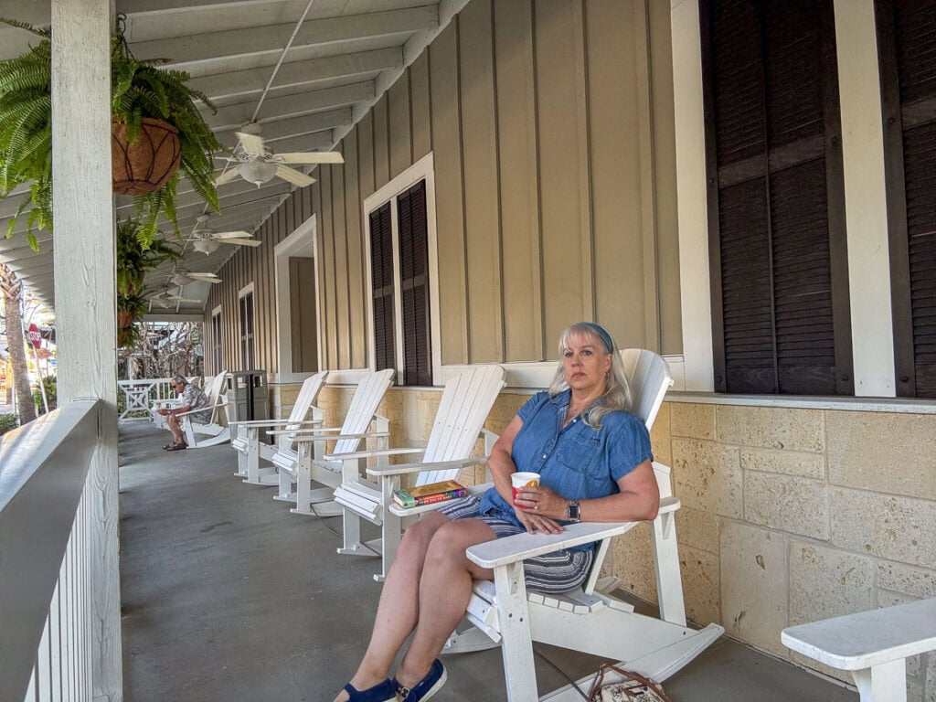 A woman in a blue dress sits on a white rocking chair on a shaded, quiet porch, holding a drink and a book—perhaps taking time to plan her new smyrna beach itinerary. Several empty chairs surround her, while potted ferns hang above, adding to the tranquil atmosphere.