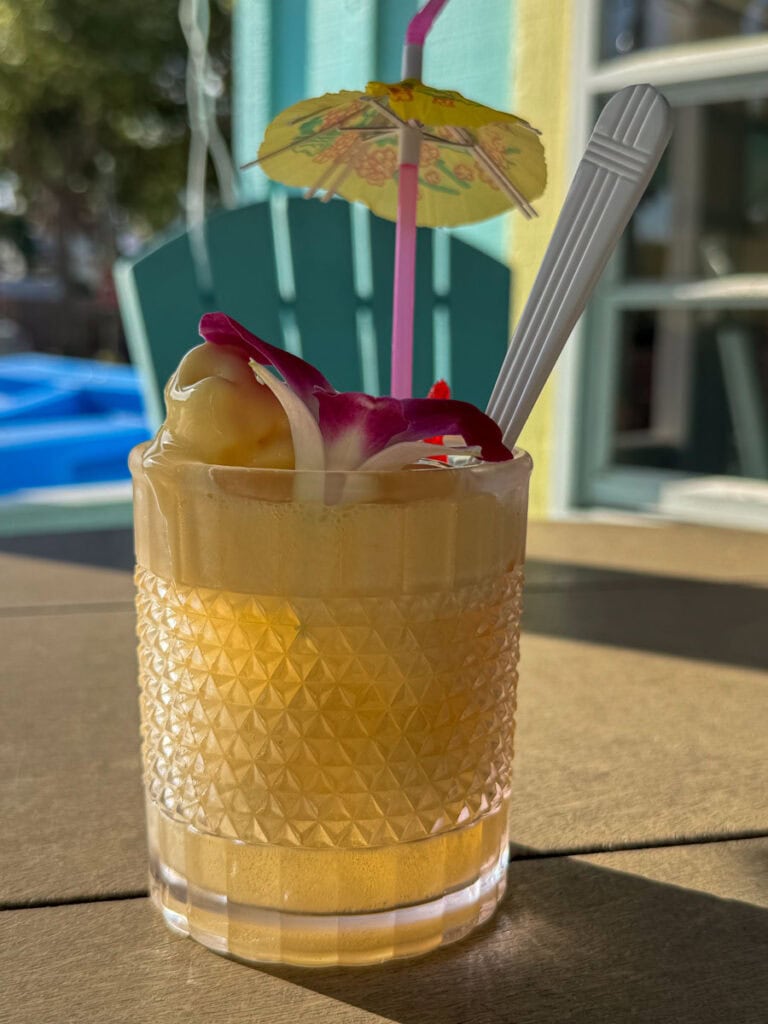 A yellow tropical drink in a textured glass with ice, garnished with a flower, fruit, a plastic spoon, and a small paper umbrella, sits on an outdoor table in sunlight—perfect for sipping during your New Smyrna Beach itinerary.