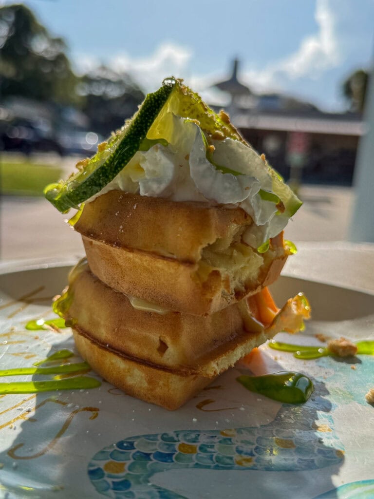 A stack of two waffles topped with whipped cream and a lime slice sits on a plate with green sauce, creating a picture-perfect breakfast that’s sure to add flavor to any New Smyrna Beach itinerary, with sunny outdoor scenery blurred in the background.