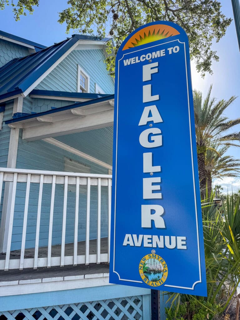Blue sign reading “Welcome to Flagler Avenue” with the New Smyrna Beach city seal, in front of a light blue building with a porch and palm trees—a perfect starting point for any New Smyrna Beach itinerary.