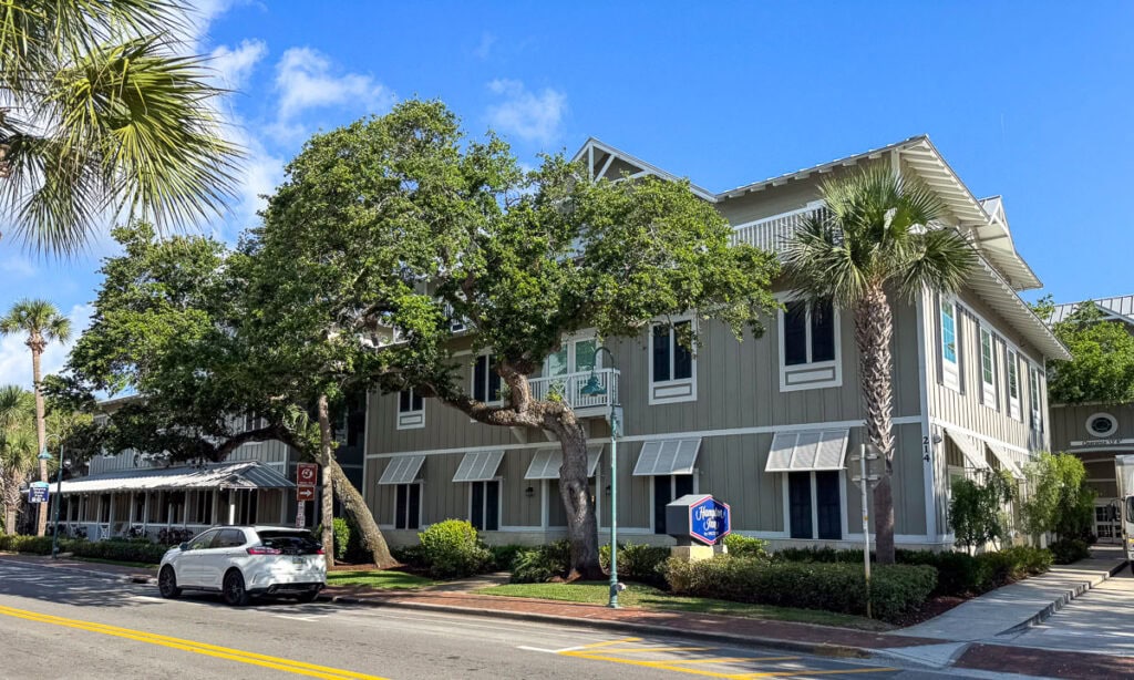A two-story gray coastal building with white trim, large windows, and awnings sits surrounded by palm trees and greenery—an inviting stop for any New Smyrna Beach itinerary. A white car is parked on the street under a bright blue sky, perfectly capturing the laid-back charm of this seaside destination.