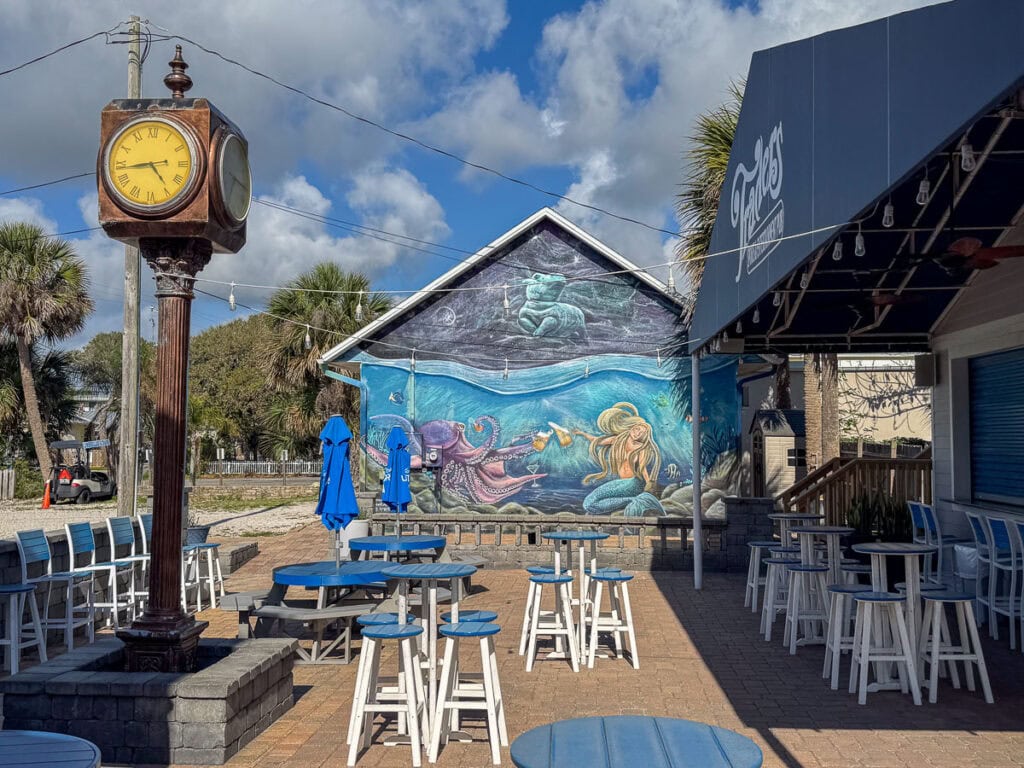 Outdoor patio with blue tables and chairs, a vintage clock, and a mural featuring an underwater mermaid scene on the wall of a small building—perfect for adding to your New Smyrna Beach itinerary. Palm trees and blue sky with clouds are visible in the background.