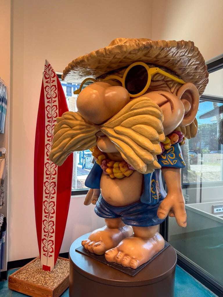 A large cartoon statue of a surfer with a big mustache, sunglasses, straw hat, Hawaiian shirt, and lei stands indoors next to a red and white surfboard, creating the perfect photo op for anyone planning their New Smyrna Beach itinerary.