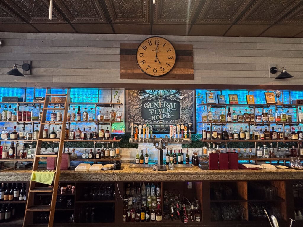 A bar with shelves of liquor bottles, a large wooden clock above, and a “General Public House” sign at the center—the perfect addition to your New Smyrna Beach itinerary. A ladder leans left, and beer taps are arranged across the marble counter.