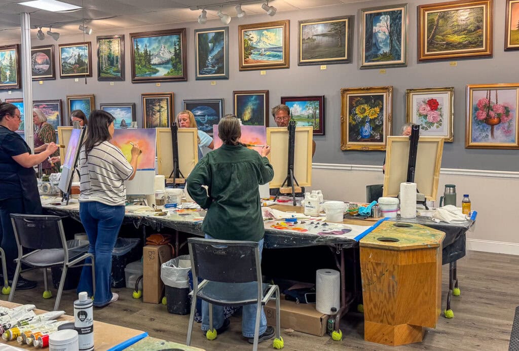 A group of people paint at easels in a brightly lit art studio, surrounded by colorful framed paintings on the walls. Art supplies and canvases are scattered across tables in the center of the room, making this creative space a must-visit stop on any New Smyrna Beach itinerary.