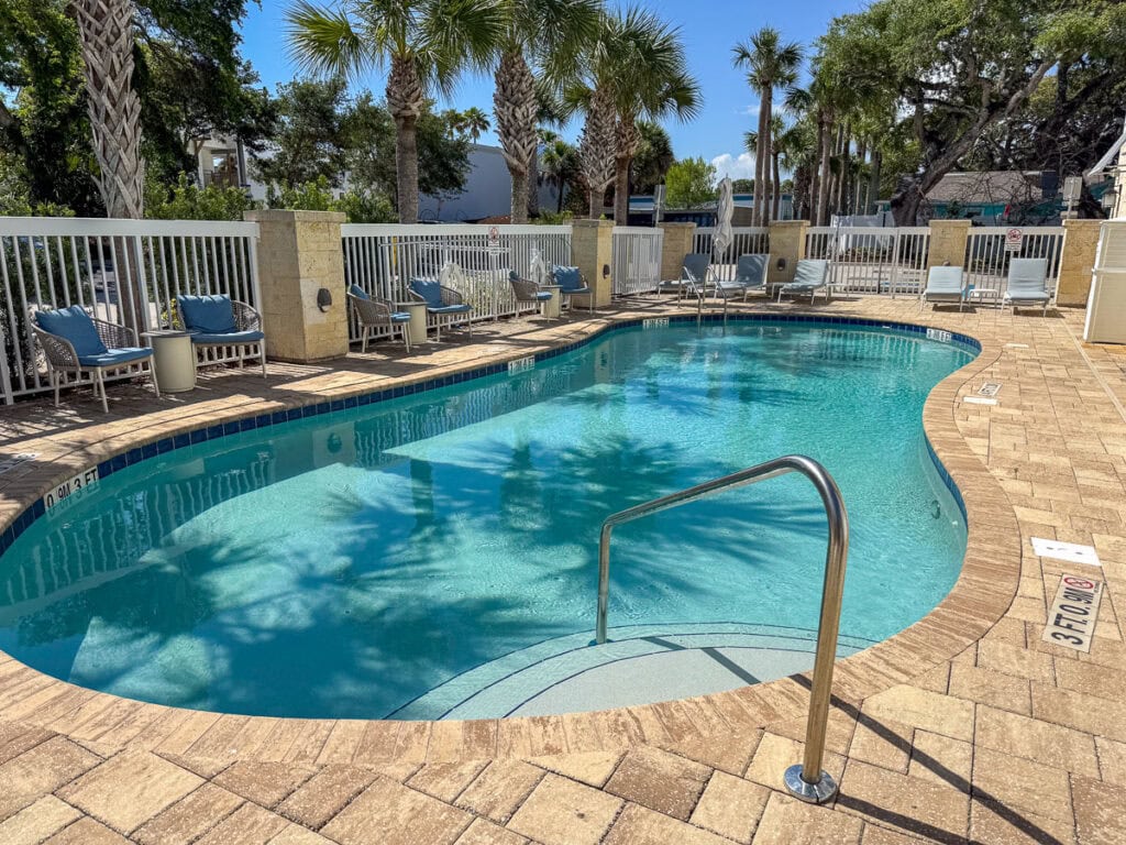 A small outdoor pool with clear water is surrounded by lounge chairs, palm trees, and a white fence on a sunny day—a perfect spot to relax during your New Smyrna Beach itinerary. A metal handrail leads into the pool.