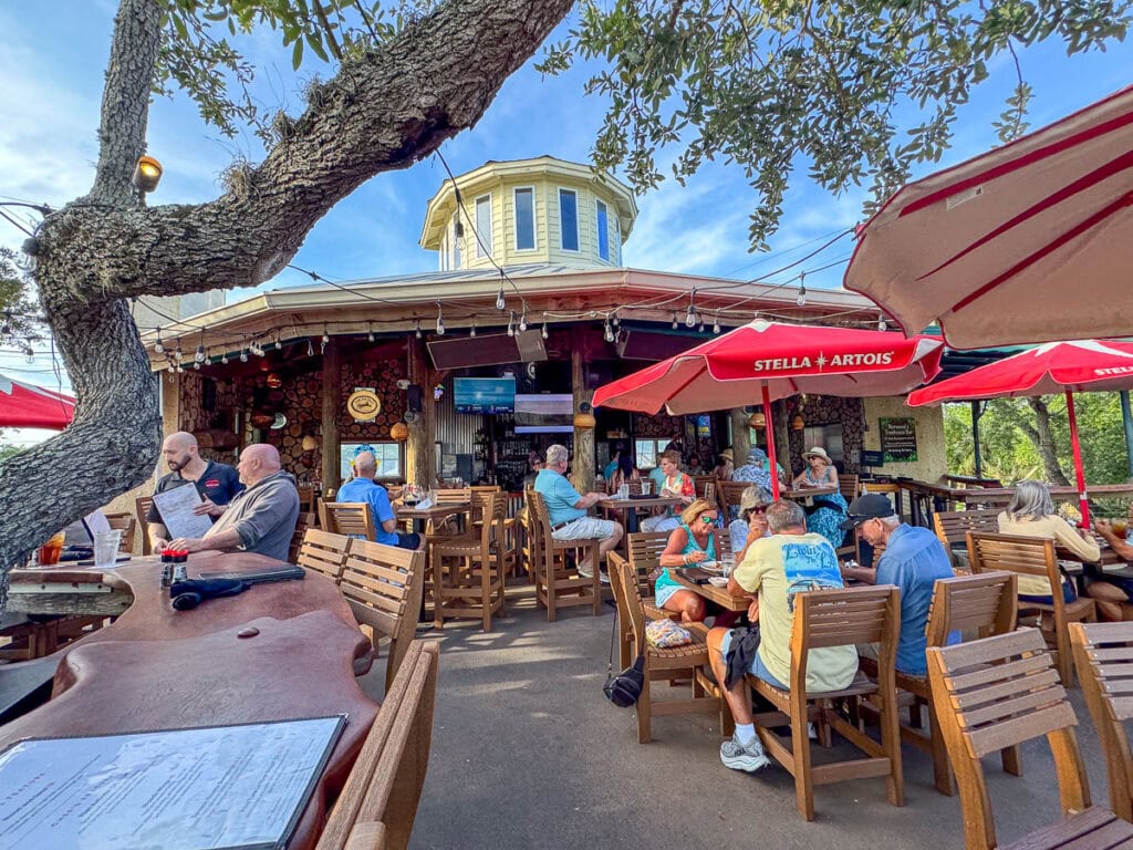 People sit at wooden tables and chairs under red umbrellas at an outdoor restaurant, making it a perfect stop on any New Smyrna Beach itinerary. A large tree extends over the seating area, offering shade and charm. The building behind them has a round upper structure and a TV above the entrance.