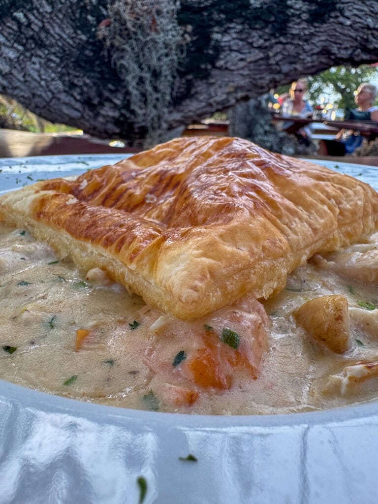 A close-up of a golden, flaky pastry sitting on creamy chicken pot pie filling, served outdoors with a large tree and people sitting at picnic tables in the background—an inviting scene that would be the perfect addition to any New Smyrna Beach itinerary.