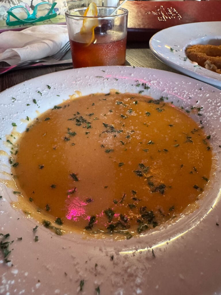 A white bowl contains orange soup garnished with green herbs—perfect for savoring on your New Smyrna Beach itinerary. A drink with ice and an orange slice sits behind it, while bright pink reflections dance on the soup’s surface. Nearby, a plate of fried food and napkins are also visible.