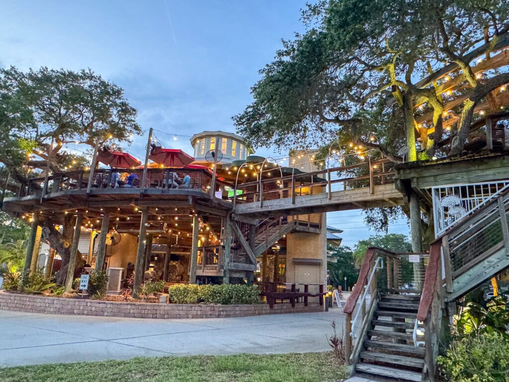 Two-story outdoor restaurant with string lights, red umbrellas, and surrounding trees at dusk—an inviting spot to add to your New Smyrna Beach itinerary. Wooden stairs lead up to a patio where people are dining under the evening sky.