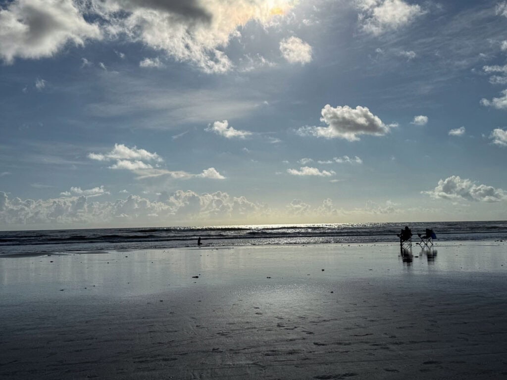 A wide sandy beach under a partly cloudy sky, with gentle waves and two empty chairs facing the ocean—an inviting start to your New Smyrna Beach itinerary. Sunlight glistens on the water, creating a calm, peaceful scene perfect for relaxation or planning your next coastal adventure.