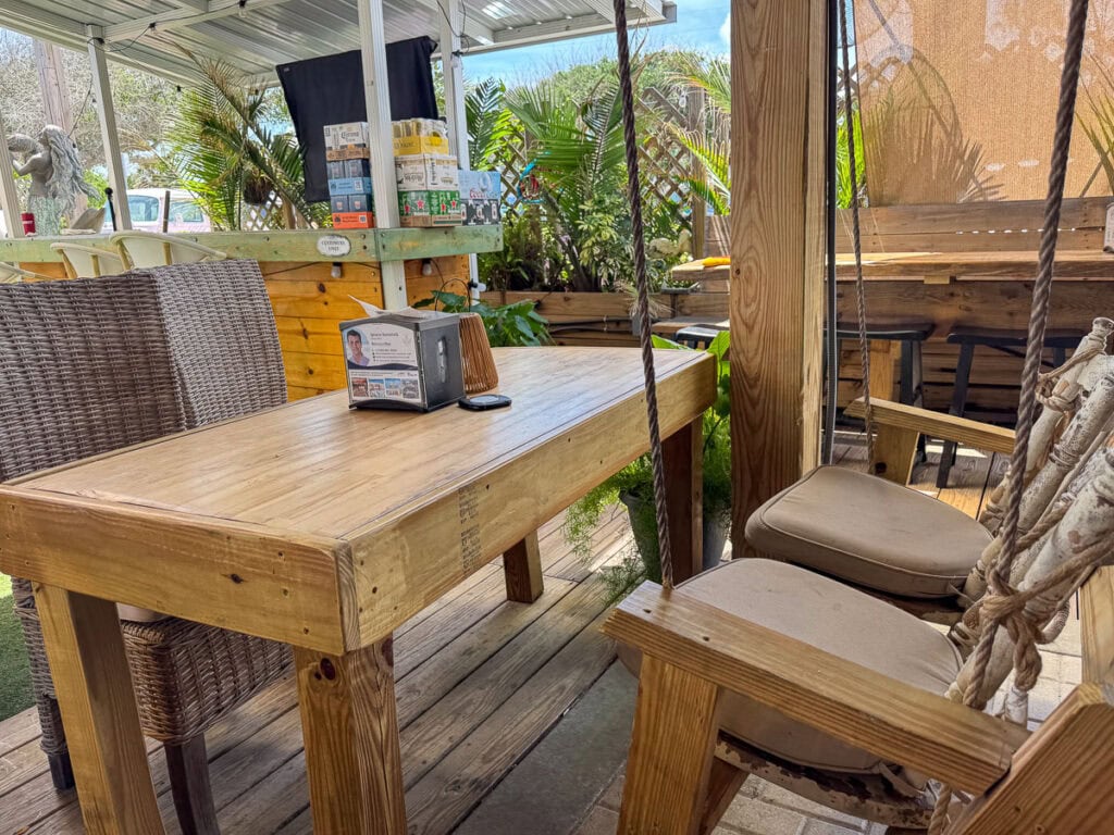 Wooden outdoor patio table with wicker and cushioned swing chairs, surrounded by plants and shaded by a canopy—just the kind of relaxing spot you’d hope to find on any New Smyrna Beach itinerary. A napkin holder sits on the table, while sunlight filters through the area, creating an inviting atmosphere.