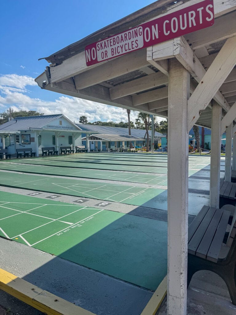 Green shuffleboard courts are shown under a covered seating area with a sign above that reads, “No Skateboarding or Bicycles on Courts.” Small buildings and palm trees are visible in the background, making this a classic stop to add to your New Smyrna Beach itinerary.
