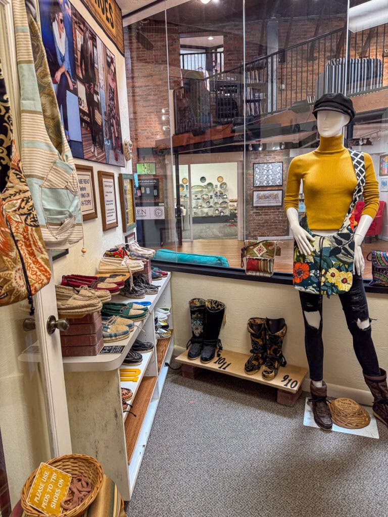 A boutique in New Smyrna Beach displays a mannequin in a yellow top, floral bag, and cap. Colorful shoes and boots line a shelf, bags hang nearby, and patterned scarves are stacked on a table. Warm lighting highlights the vintage décor—making it a charming stop to add to your New Smyrna Beach itinerary.