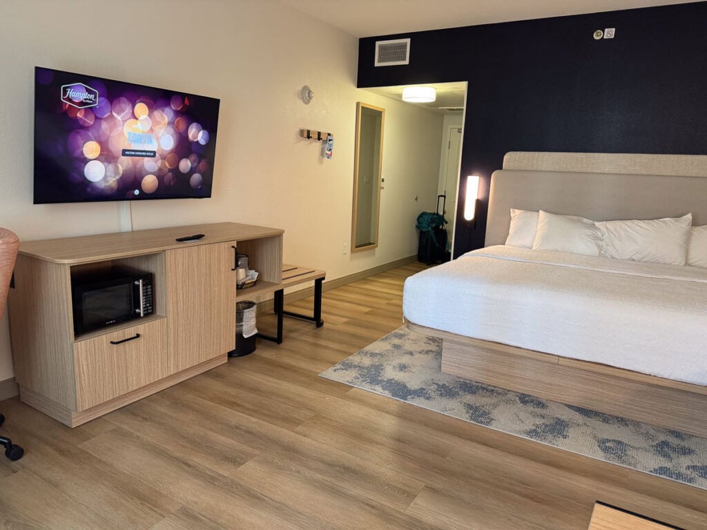 Modern hotel room with a large bed, wall-mounted TV showing a screensaver, wooden furniture, a microwave, and a mini fridge—perfect for relaxing after exploring your New Smyrna Beach itinerary. There’s a mirror and coat hooks near the entrance, and the room features light wood flooring for a bright, welcoming atmosphere.