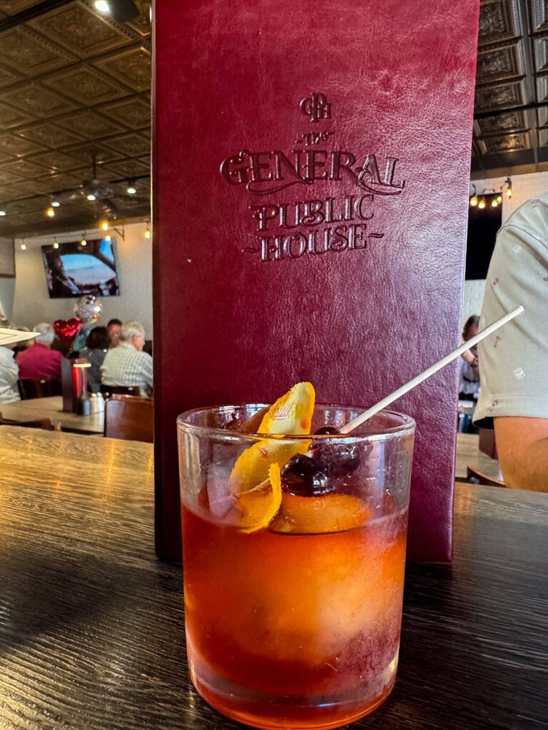 A glass of an Old Fashioned cocktail garnished with an orange peel and cherry sits on a wooden bar, with a maroon “General Public House” menu in the background—an inviting stop to consider for your New Smyrna Beach itinerary.