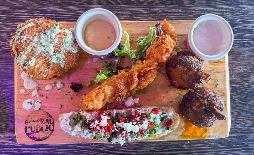 A wooden board with a burger, fried fish fillet, salad-topped bread, two stuffed mushrooms, and two dipping sauces—each element ready to elevate your New Smyrna Beach itinerary. “General Public” is stamped on the board.