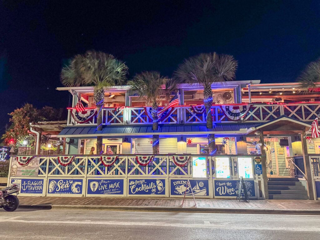 A brightly lit seaside tavern at night, decorated with flags, palm trees, and banners. The signs advertise cocktails, live music, and seafood. With its welcoming, festive atmosphere, this lively spot is a perfect addition to any New Smyrna Beach itinerary.