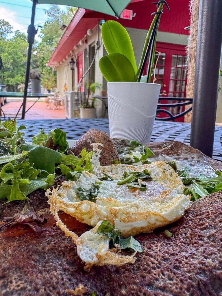 A close-up of a fried egg on a brown crepe with greens, on a metal table outdoors—perfect for fueling up before your New Smyrna Beach itinerary. In the background, a white potted plant and patio seating rest under a green umbrella, capturing the relaxed atmosphere of your coastal adventure.