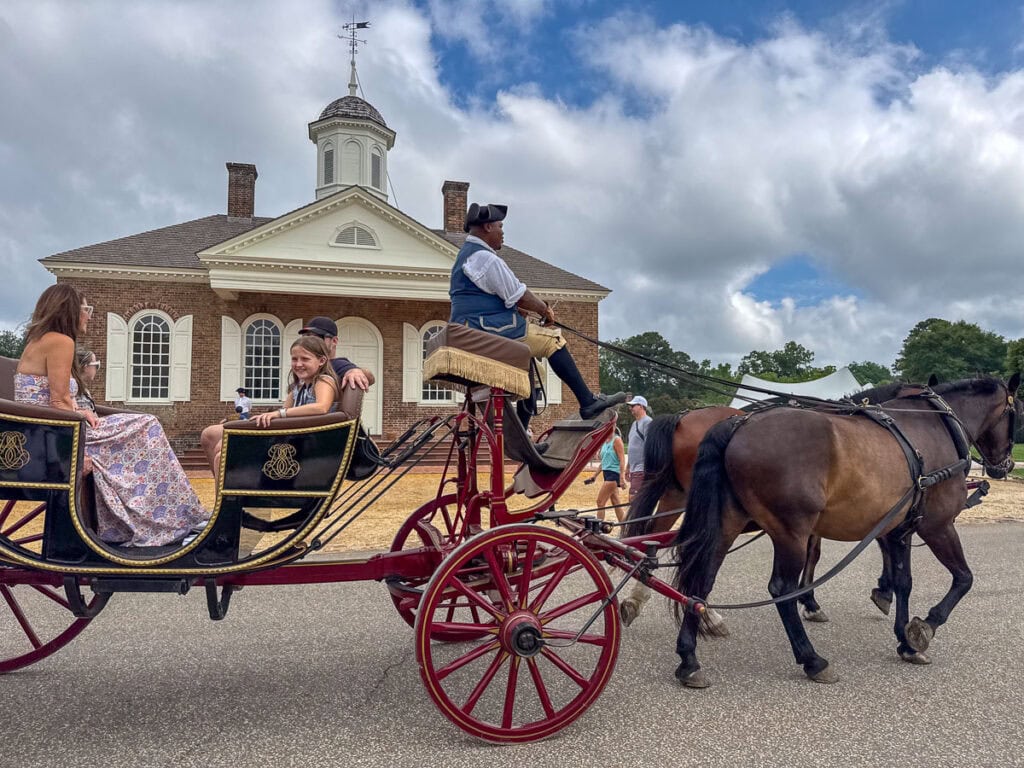 A horse-drawn carriage with a driver and three passengers passes by a historic brick building with a white cupola under a partly cloudy sky, offering one of the many classic family activities and things to do in Williamsburg.