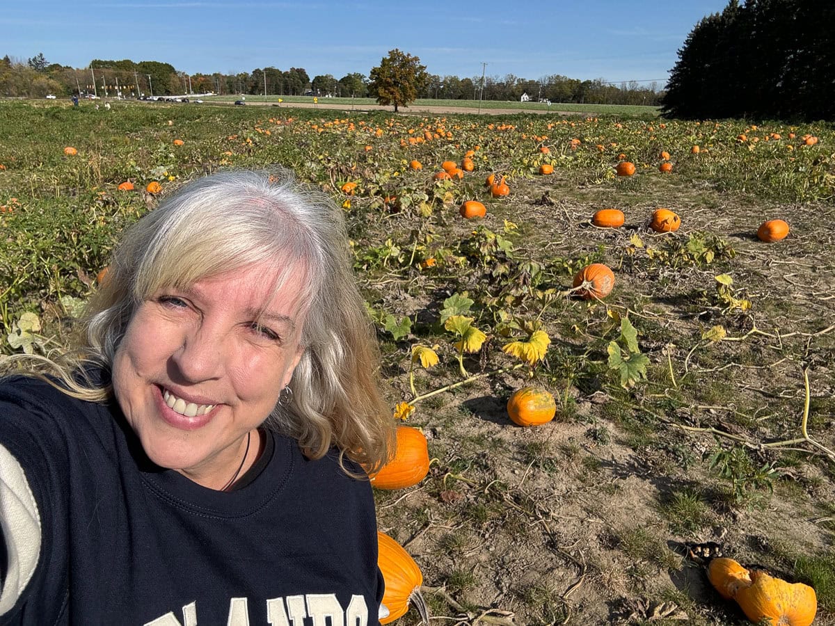 A smiling woman with gray hair takes a selfie in a sunny pumpkin patch, surrounded by scattered orange pumpkins and green vines—a perfect example of Fall Things to do in the Ann Arbor area.
