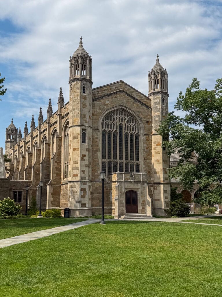 Gothic-style stone building with large arched windows and two towers, set on a green lawn under a partly cloudy sky—an impressive sight to experience during a multigenerational trip to Ann Arbor.