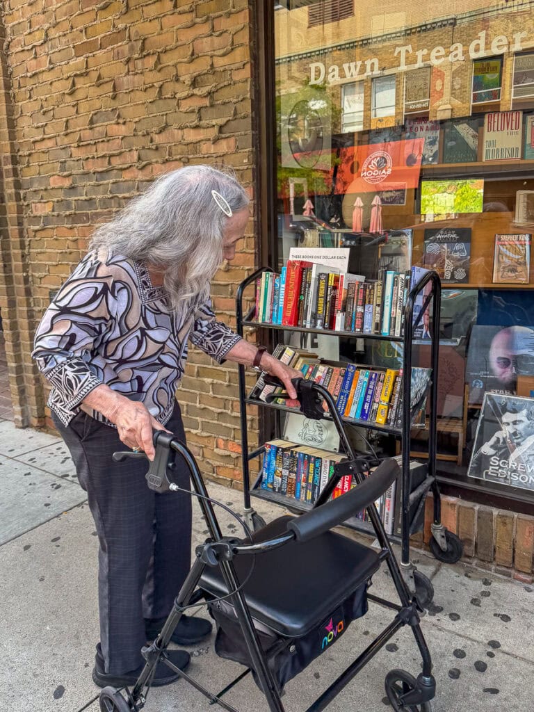 An elderly woman with gray hair and a walker browses books on a cart outside a bookstore with a brick wall and glass windows displaying books in the background, enjoying a quiet moment during her multigenerational trip to Ann Arbor.