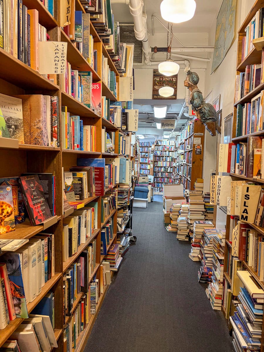 A narrow aisle in a bookstore lined with tall shelves packed with books beckons visitors to explore, much like discovering new Fall Things to do in the Ann Arbor. Stacks of books sit on the floor, and a chair is visible in the well-lit background. Signs label book genres along the shelves, inviting readers to linger and enjoy a cozy afternoon indoors.