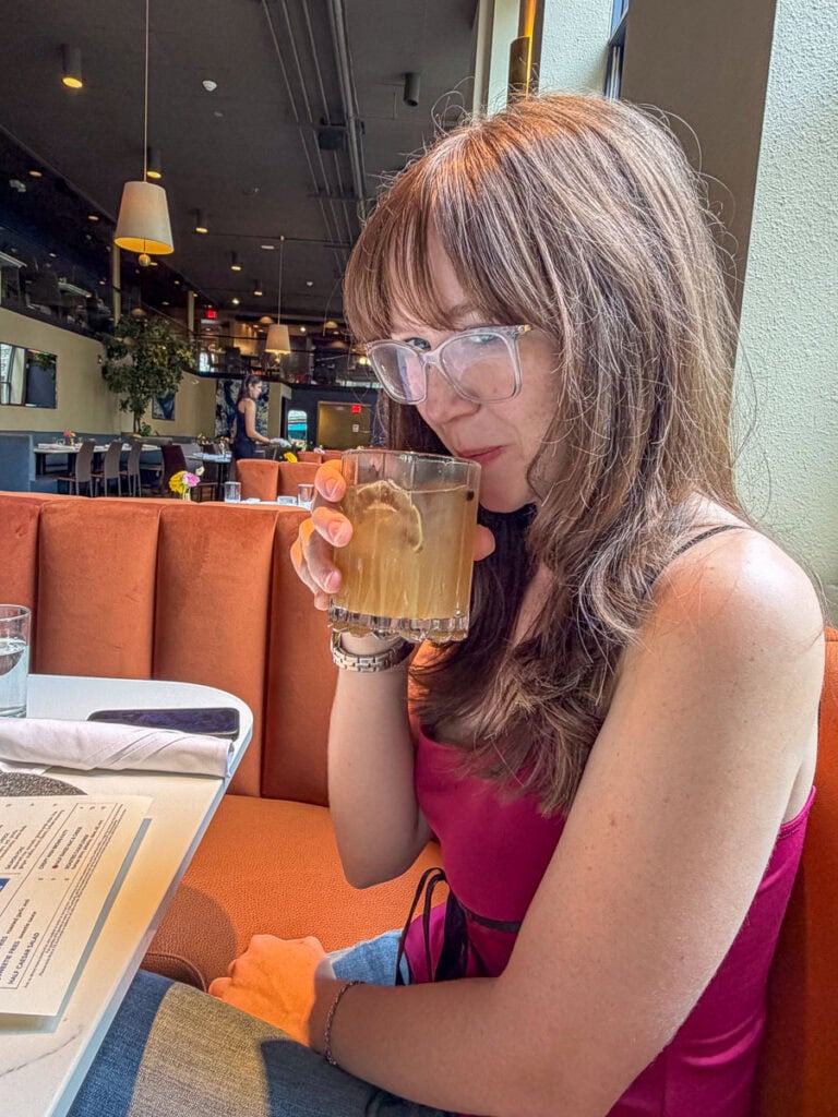 A woman with long brown hair and glasses sips a cocktail at a restaurant booth. She wears a sleeveless magenta top and sits beside a menu, taking in the modern restaurant setting after visiting your kid at the University of Michigan.