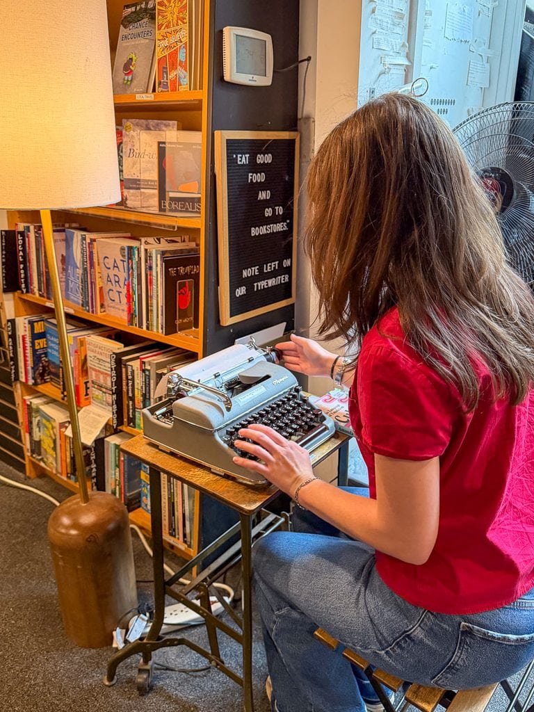 A person in a red shirt types on a vintage typewriter in a bookstore, surrounded by bookshelves. A sign nearby reads, “Eat good food and go to bookstores. Note left on our typewriter.” This cozy scene is the perfect inspiration for your list of Fall Things to do in the Ann Arbor area.