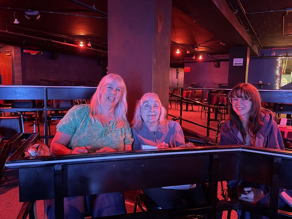 Three women sit and smile at a table inside a dimly lit room with red lighting, empty seats, and tables around them, capturing a warm moment from their multigenerational trip to Ann Arbor.