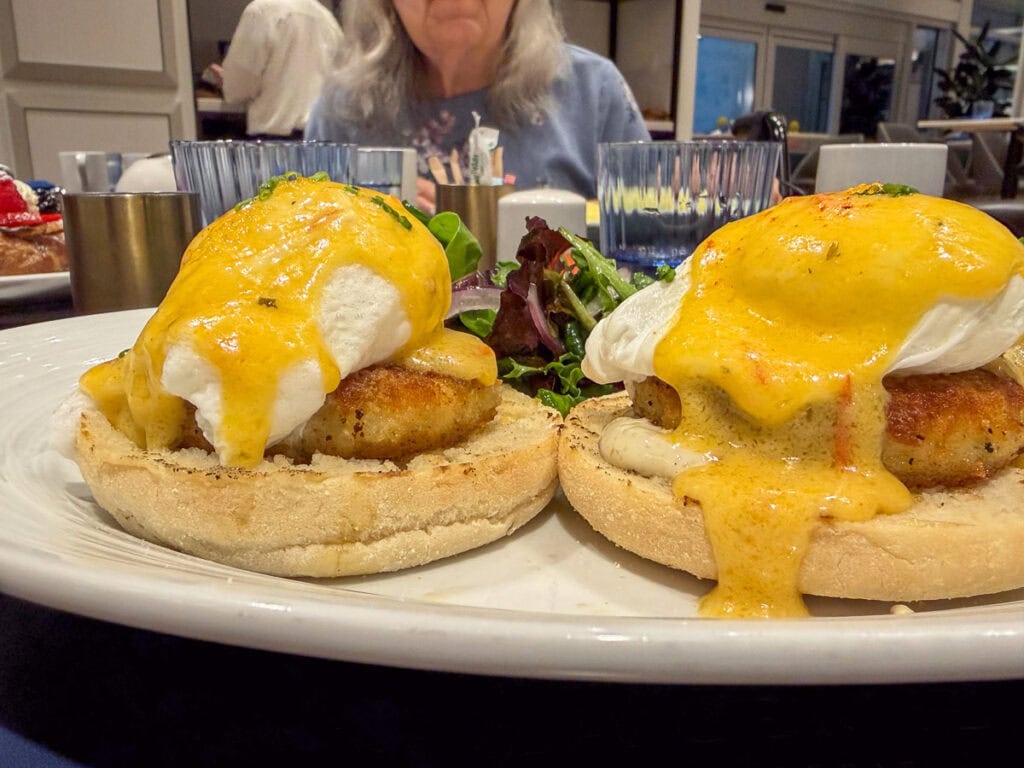 Two poached eggs with hollandaise sauce on crab cakes and English muffins, served with greens on a white plate; perfect for anyone visiting the University of Michigan, while a person is blurred in the background.