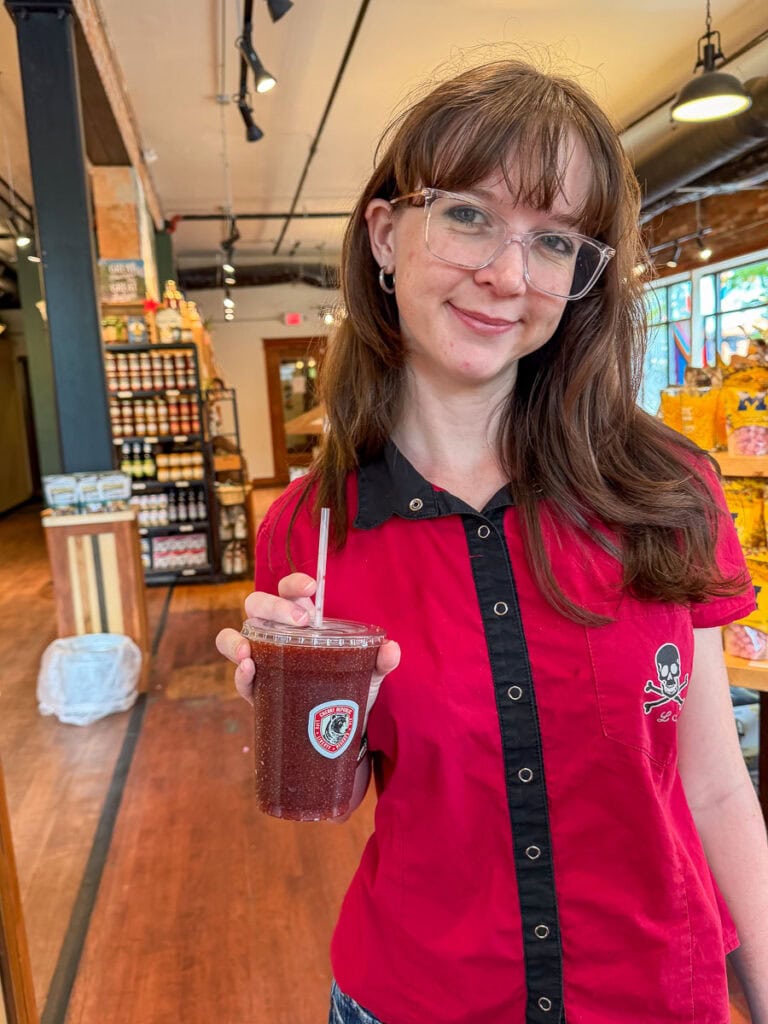 Woman with glasses in a red shirt holds a dark smoothie in a plastic cup, standing in a shop with wooden floors and shelves of products in the background—perhaps taking a refreshing break during her multigenerational trip to Ann Arbor.