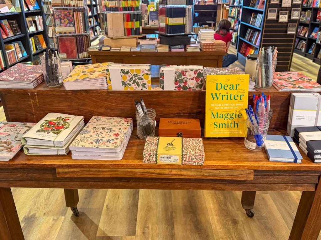 A wooden table in a bookstore displays floral notebooks, pens in jars, and the yellow book “Dear Writer” by Maggie Smith—an inviting scene that could easily be discovered on a multigenerational trip to Ann Arbor. Shelves full of books and a person sitting are visible in the background.