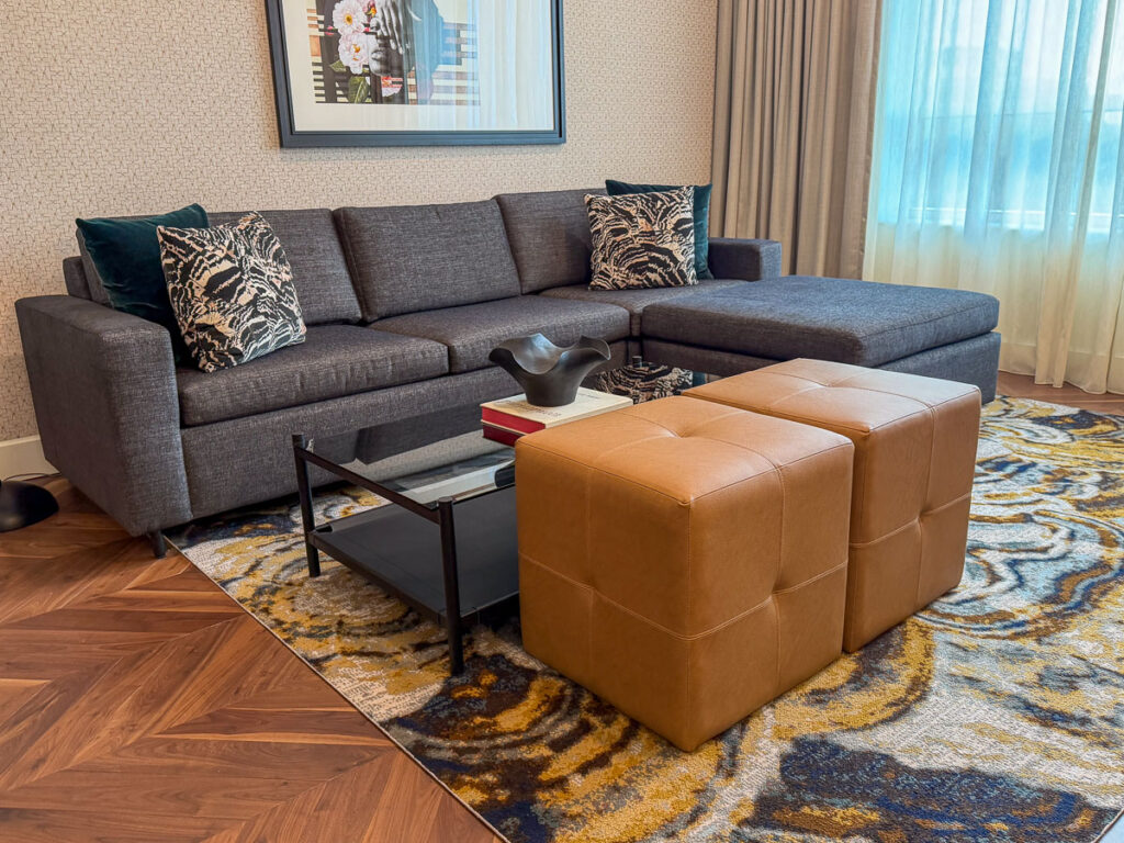 A modern living room with a gray sectional sofa, patterned pillows, two tan ottomans, a glass coffee table, and a colorful rug on wood floors—perfect for relaxing after a multigenerational trip to Ann Arbor. A framed artwork hangs above the sofa near large windows with light curtains.