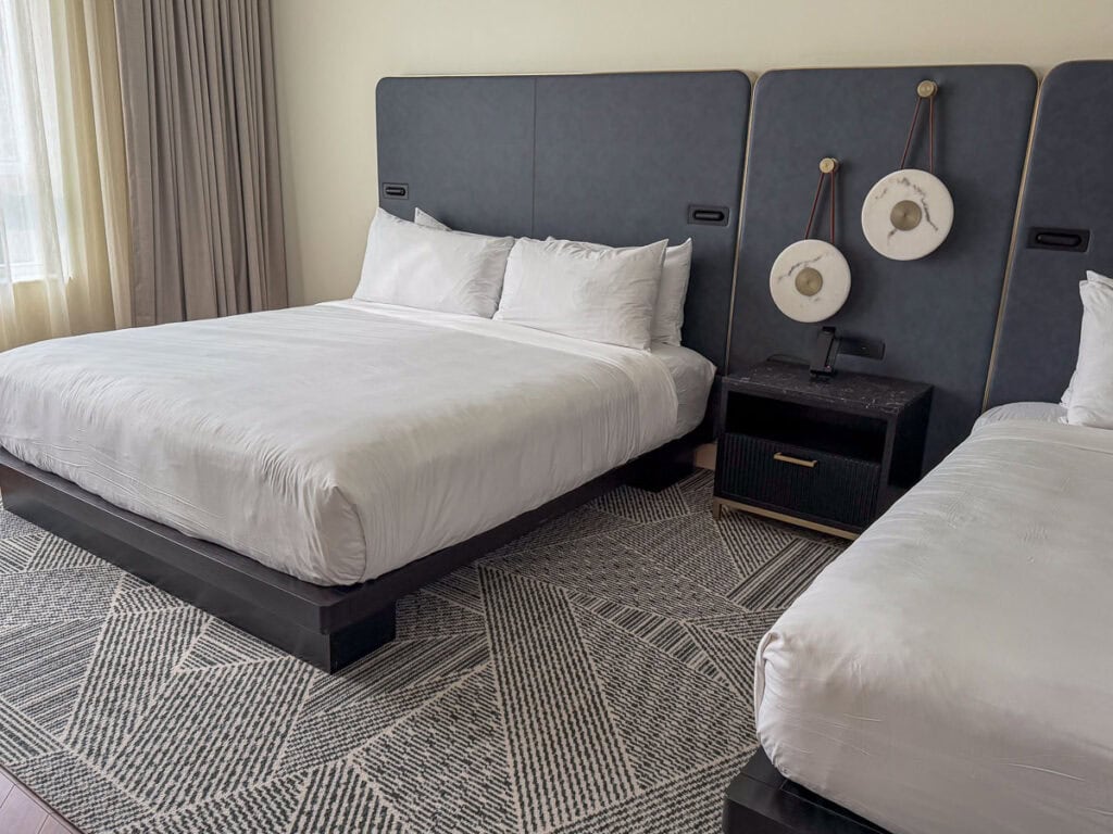 A hotel room with a large bed and a smaller bed, both with white bedding. There’s a black nightstand between them and circular wall décor above the headboards. A patterned rug covers the floor. Light enters from a window, making it an inviting retreat after enjoying fall things to do in the Ann Arbor area.
