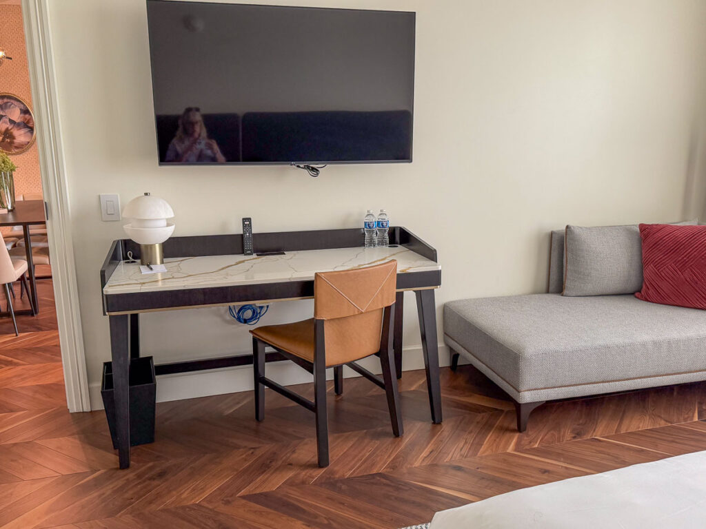 A modern hotel room, perfect for a multigenerational trip to Ann Arbor, features a desk with a chair, lamp, two water bottles, TV mounted above, and a gray chaise lounge with a red pillow. A reflection of a person is visible in the TV screen.