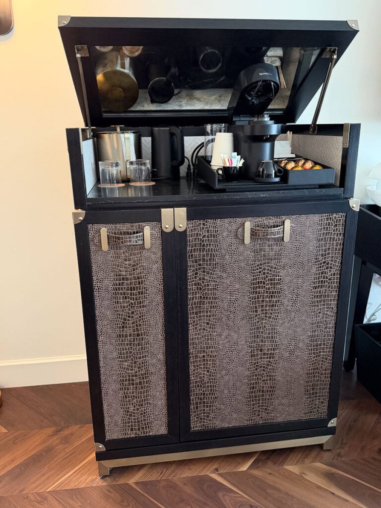 A minibar cabinet with a textured front holds a coffee machine, cups, glasses, and condiments on top—perfect for cozying up after exploring Fall Things to do in the Ann Arbor area. The lid is open, revealing inside storage and coffee supplies for your seasonal refreshments.