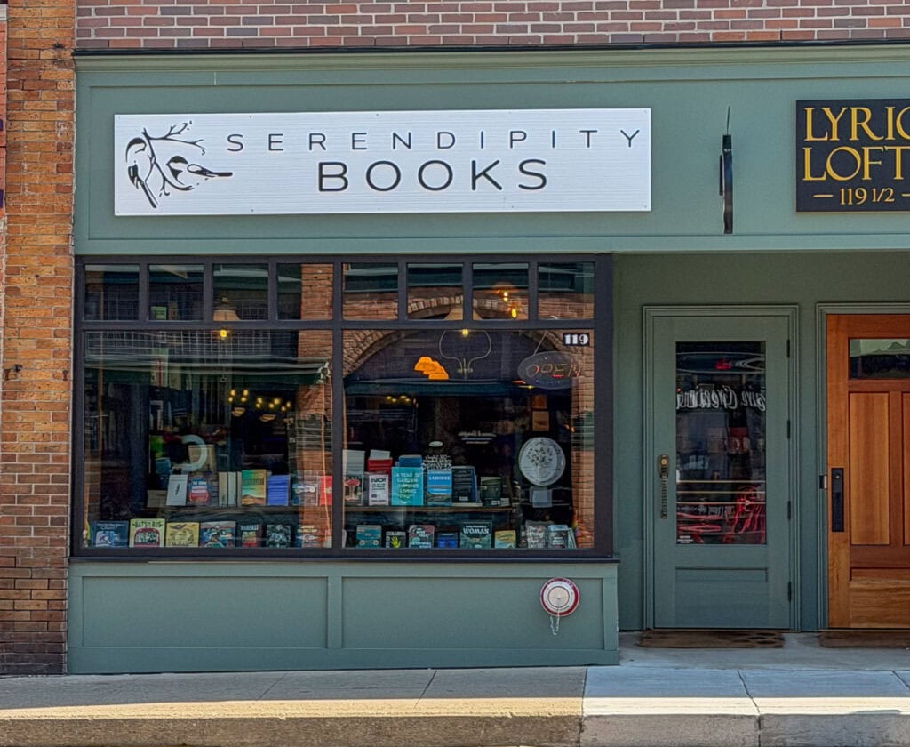 Storefront of Serendipity Books with large display windows showing books inside—a perfect stop during a multigenerational trip to Ann Arbor. The sign above the window has a simple branch design. Next door is Lyric Loft, with a green door and gold lettering.