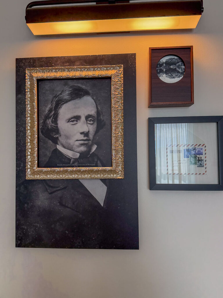 A wall features a framed black-and-white portrait of a man in a suit, a small framed photo of the moon, and a framed envelope with stamps—keepsakes from a memorable multigenerational trip to Ann Arbor—all lit by a wall-mounted light above.