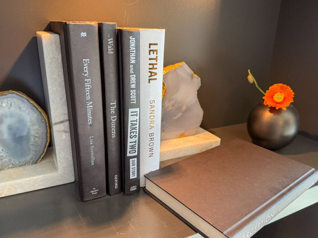 Books stand upright between two geode bookends on a shelf, hinting at cozy afternoons spent reading and planning Fall Things to do in the Ann Arbor area. A closed notebook lies nearby, ready to capture new ideas or autumn adventures. A small black vase with an orange flower decorates the scene, set against a plain wall for a warm and inviting seasonal touch.