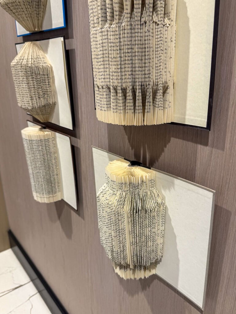 Four old books with pages intricately folded into geometric shapes are mounted on a wooden wall, each displayed on a white rectangular background as part of an art installation—an inspiring showcase that’s perfect for your list of fall things to do in the Ann Arbor area.
