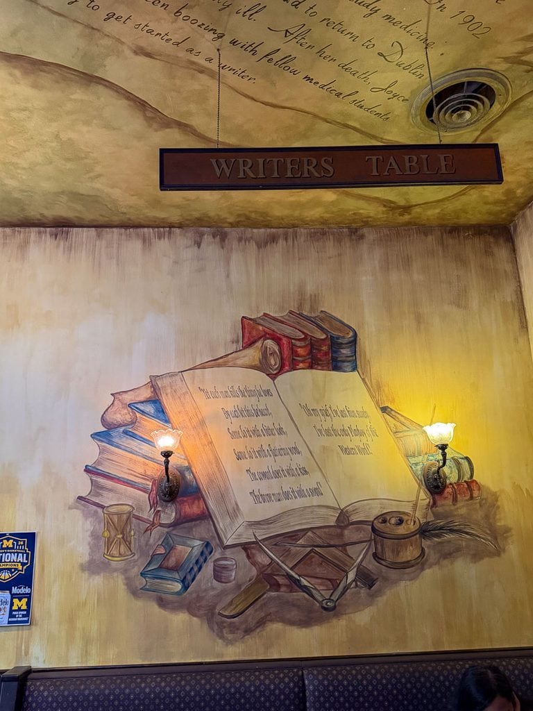 A mural of open books, ink, and candles is painted on a wall beneath a “Writers Table” sign, with warm lighting and a decorative script on the ceiling above—an inviting scene reminiscent of the creative energy you might discover during a multigenerational trip to Ann Arbor.