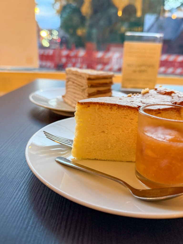 A close-up of a white plate with a slice of sponge cake and a glass of orange sauce, with a fork—perfect for savoring after exploring fall things to do in the Ann Arbor area. In the background, another plate has a layered dessert, and a candle is visible on the table.