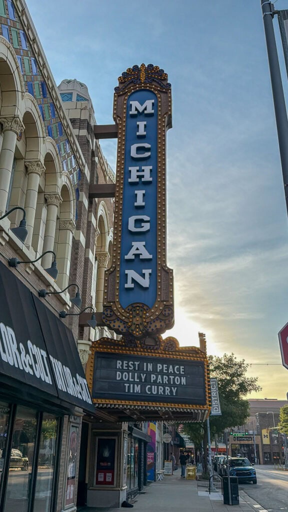 Vertical Michigan theater sign on a city street. Marquee below reads: “REST IN PEACE DOLLY PARTON TIM CURRY.” The sun is setting behind the building, casting a warm glow perfect for a multigenerational trip to Ann Arbor. Storefronts line the sidewalk.
