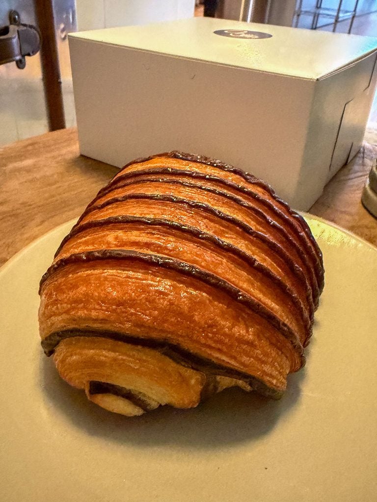 A pain au chocolat pastry with chocolate stripes sits on a plate, evoking the comforting memories of a multigenerational trip to Ann Arbor, with a white box and wooden table in the background.