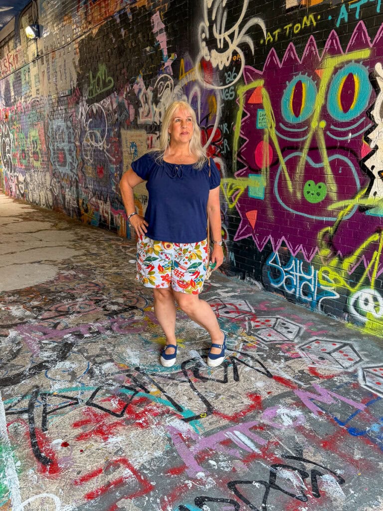 A woman stands confidently in a graffiti-covered alley while visiting the University of Michigan, wearing a navy shirt, colorful shorts, and blue shoes. Bright street art and painted designs fill the wall and ground around her—a vibrant scene that could be featured in any parent’s guide to exploring campus life.