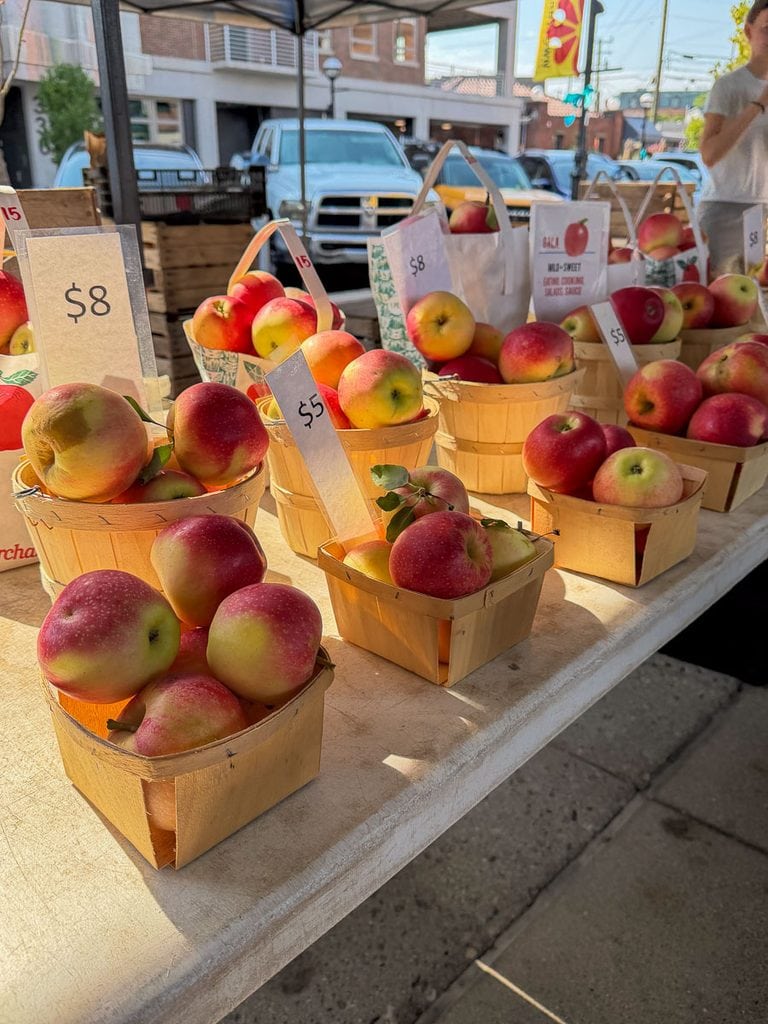 Small wooden baskets filled with red and yellow apples are displayed on a market table with price tags of $5 and $8, creating a charming scene perfect for a multigenerational trip to Ann Arbor. Cars and buildings are visible in the background.
