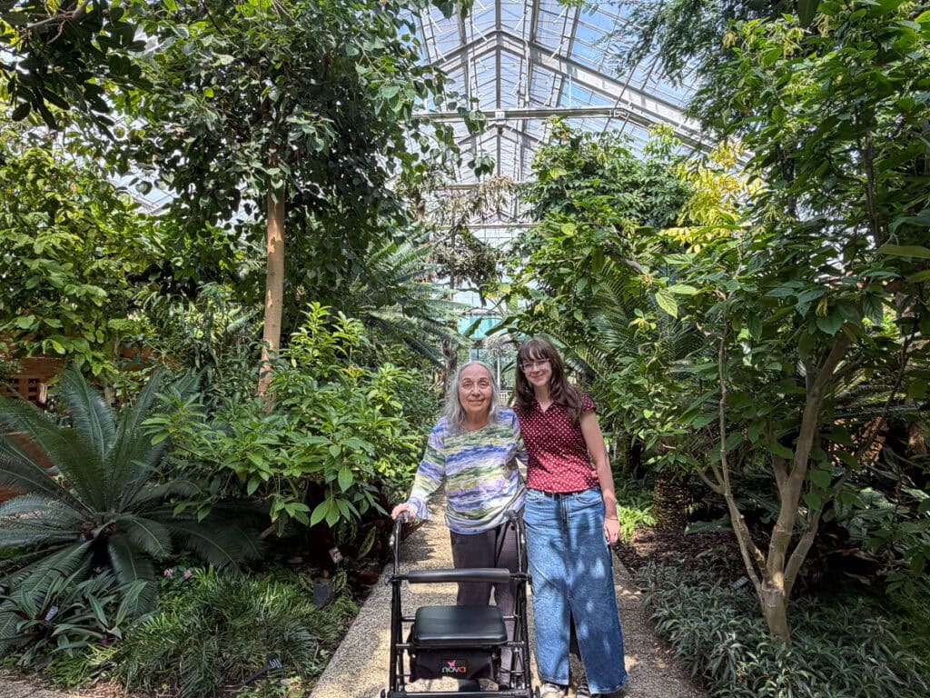 Two women, one with a walker, stand smiling together on a gravel path surrounded by lush green plants in a large, sunlit greenhouse—capturing a special moment from their multigenerational trip to Ann Arbor.