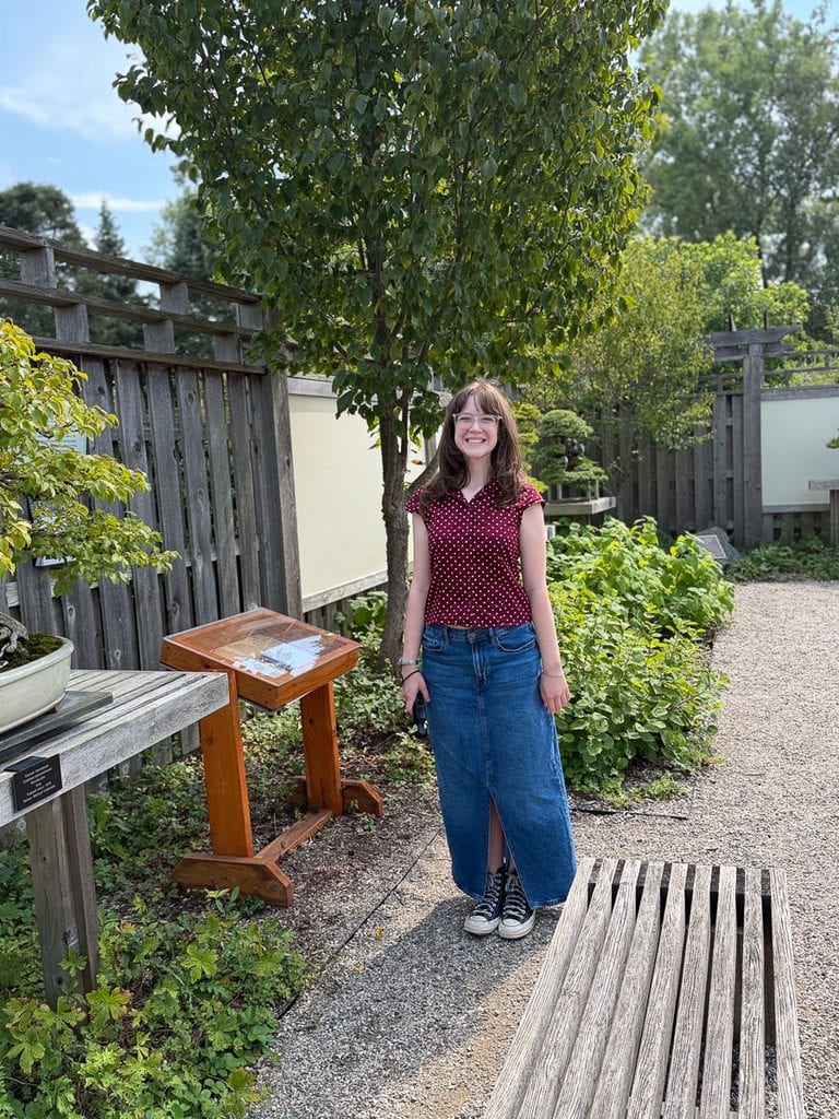 A smiling girl with glasses stands on a gravel path in a garden, near a wooden bench and an informational sign, wearing a red polka dot top, long denim skirt, and black sneakers—a perfect moment reminiscent of visiting your kid at the University of Michigan.